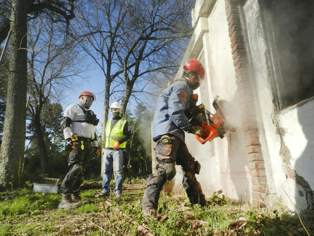 Construction workers dressed in safety gear demolishing a brick wall with heavy machinery outdoors.